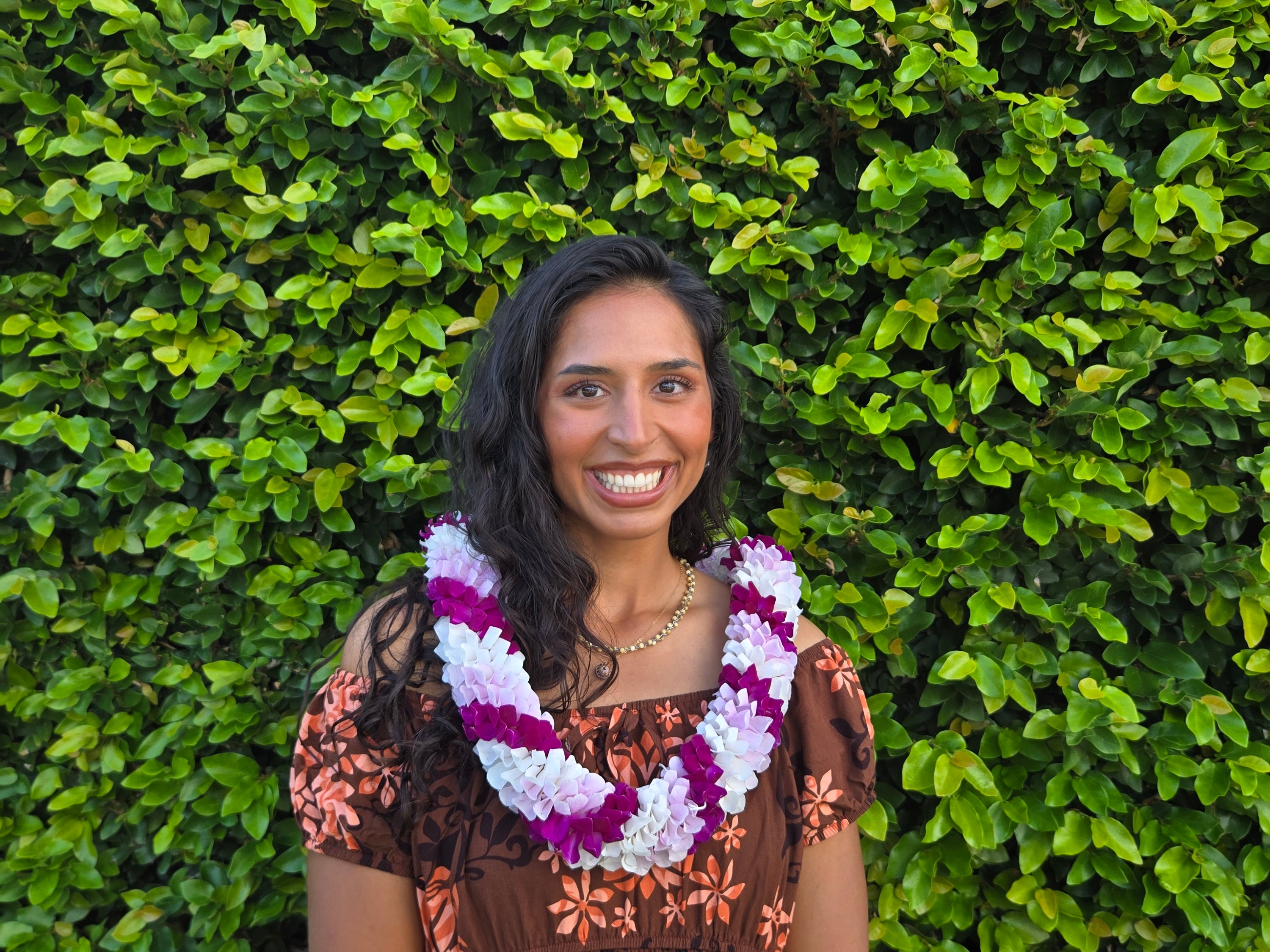Woman wearing a handcrafted white, purple, and pink spiral orchid lei in Hawaii, standing in front of lush green foliage.